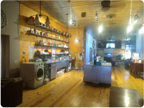 A spacious interior with wooden floors, shelves of items, a washing machine, and a cozy seating area in the background.