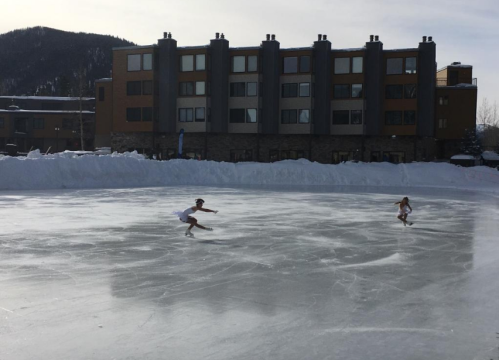 Two figure skaters perform on an outdoor ice rink, with snow and buildings in the background.