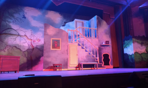 A colorful stage set featuring a staircase, door, and painted backdrop of trees and mountains, illuminated by stage lights.