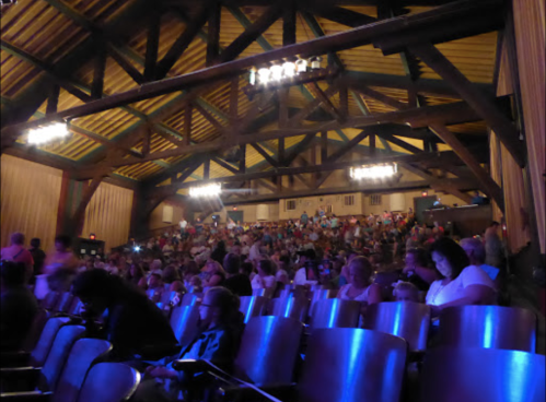 A crowded theater with wooden beams and purple lighting, showing an audience settling in for a performance.