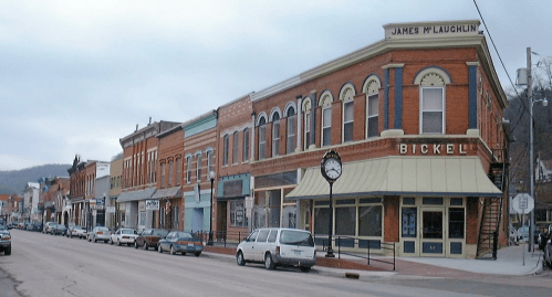 Historic brick buildings line a street, featuring shops and a clock, with parked cars along the curb.