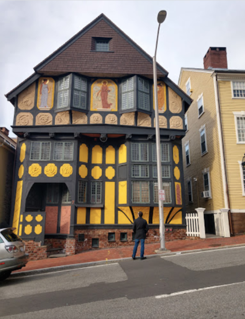 A person stands in front of a colorful, ornate building with yellow and black patterns and decorative murals.