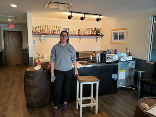 A woman stands in a cozy bar area with a wooden barrel and shelves of bottles in the background.