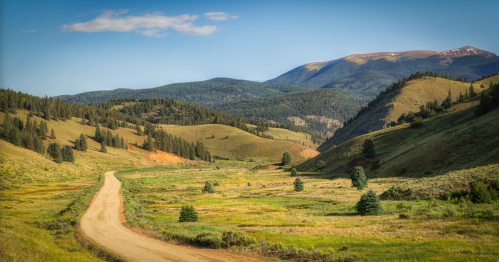 A winding dirt road through lush green hills and mountains under a clear blue sky.