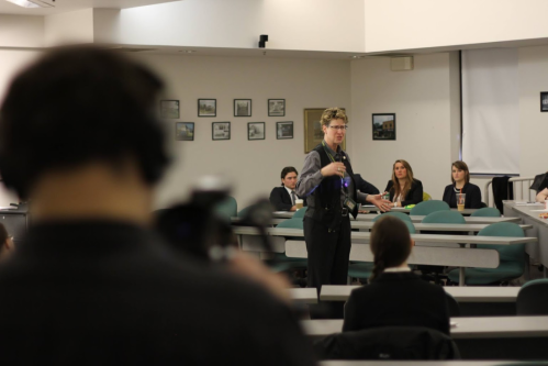 A speaker presents to an audience in a conference room, with a camera operator in the foreground.