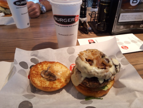 A burger with melted cheese, sautéed onions, and mushrooms on a bun, next to a drink from Burger 21.
