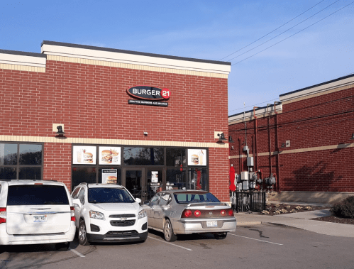 Exterior of a Burger 21 restaurant with a brick facade, parked cars in front, and clear blue sky above.