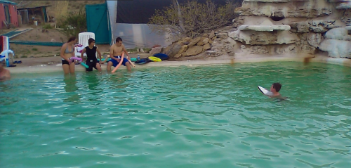 A group of children relax by a green pool, while one swims and another holds a float. Rocks and a building are in the background.