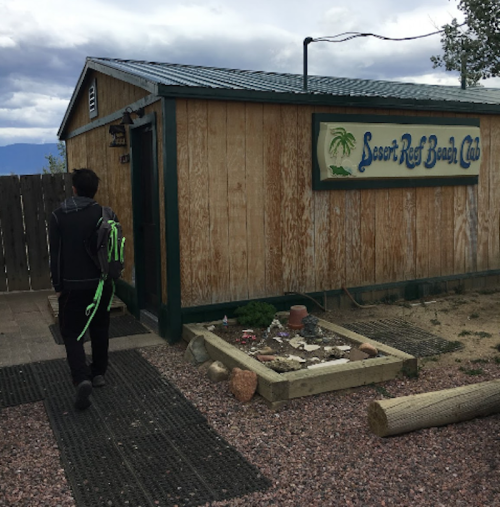 A person walks towards a wooden building with a sign that reads "Desert Reef Beach Club" under a cloudy sky.