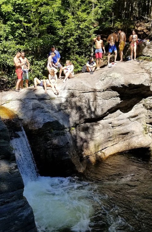 A group of people relax by a rocky riverbank, enjoying a sunny day near a small waterfall and swimming area.