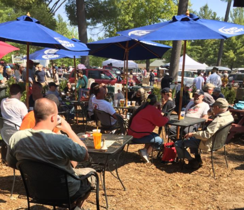 A lively outdoor gathering with people sitting at tables under blue umbrellas, enjoying drinks and conversation.