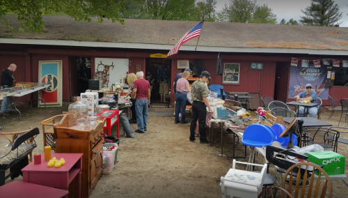 A bustling outdoor flea market with various items for sale, people browsing, and an American flag in the background.