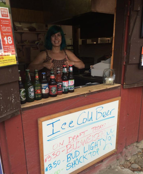 A woman with teal hair stands behind a bar, smiling, with various beer bottles displayed and a sign advertising prices.