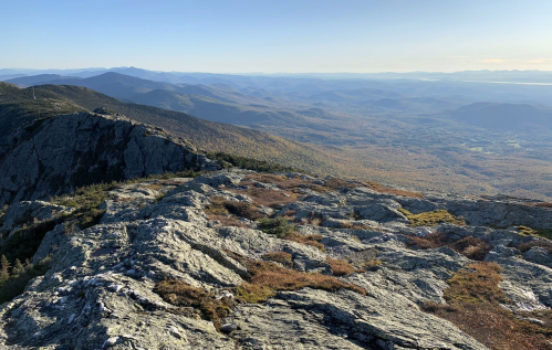 A panoramic view from a rocky mountain peak, showcasing rolling hills and valleys in autumn colors under a clear sky.