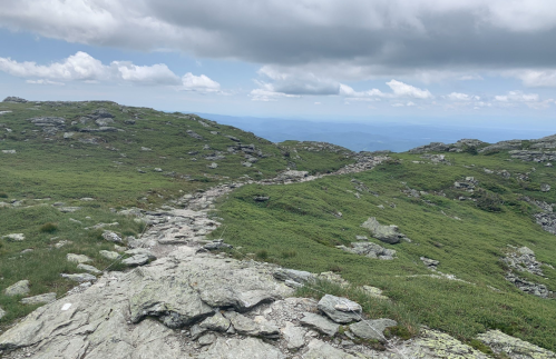 A rocky trail winds through green hills under a cloudy sky, with distant mountains visible in the background.