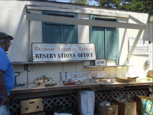 A man stands by a wooden stall with an "Outdoor Antiques Market Reservations Office" sign and various antiques displayed.