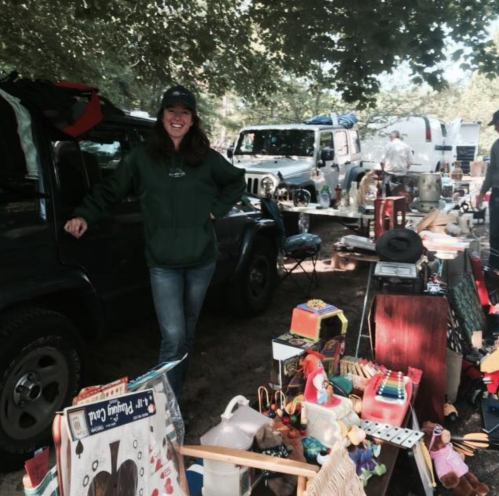 A woman in a green hoodie stands beside a table of assorted items at a flea market, with vehicles in the background.