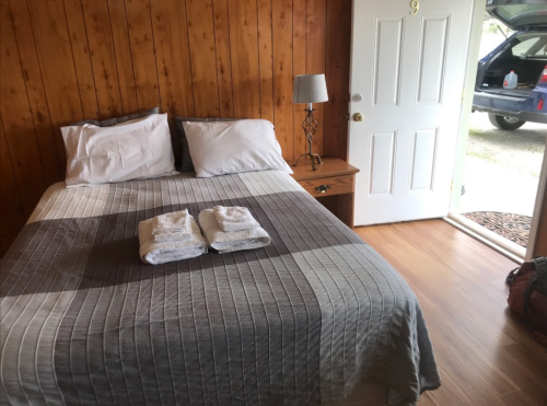 Cozy bedroom with a double bed, fresh towels, wooden walls, and an open door leading outside.