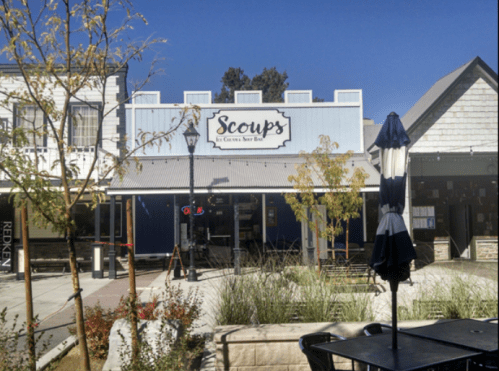 A storefront with the sign "Scoops" in a sunny outdoor setting, featuring trees and a blue sky.