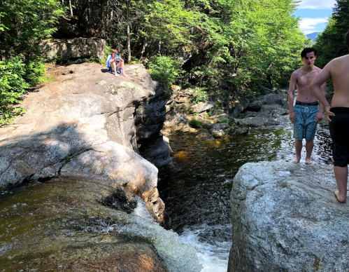 Two people near a rocky riverbank, one sitting on a rock and another standing by the water, surrounded by greenery.