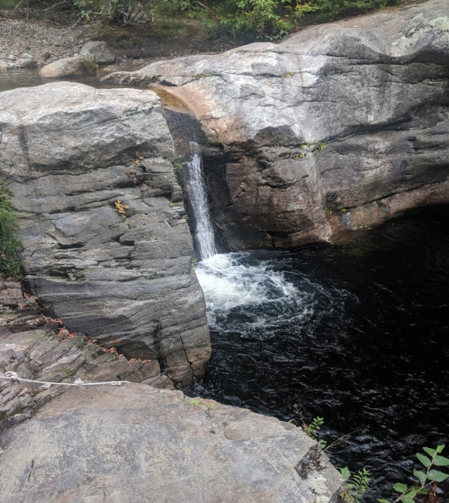 A small waterfall cascades over rocks into a dark pool surrounded by greenery.