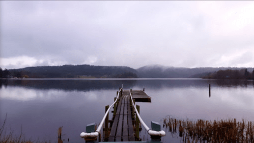 A serene lake view with a wooden dock extending into calm waters, surrounded by misty hills and overcast skies.