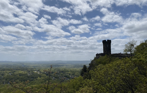 A stone tower on a rocky outcrop overlooks a lush green valley under a partly cloudy sky.