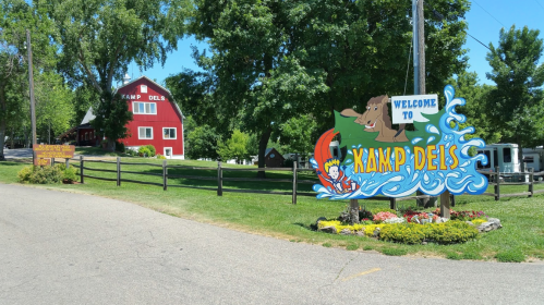 Colorful sign welcoming visitors to Kamp Dels, with a red barn and lush greenery in the background.