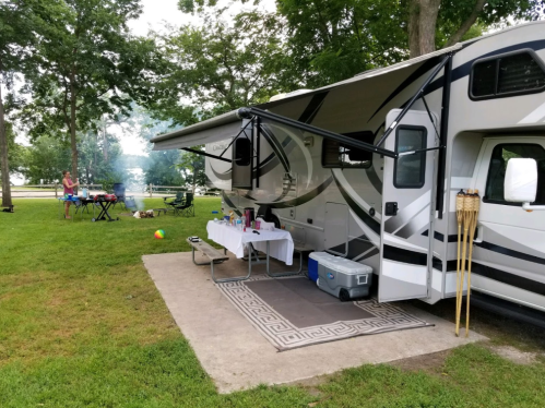 A parked RV with an awning, outdoor table, and cooking setup in a grassy area surrounded by trees.