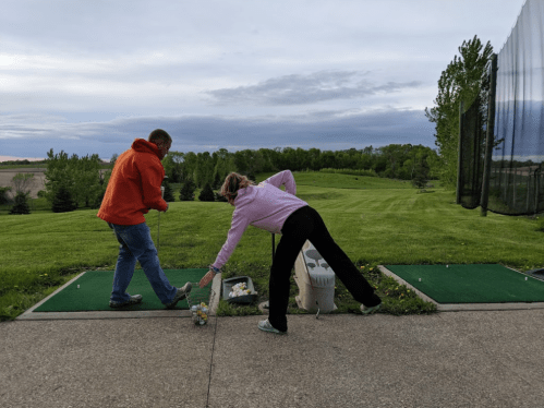 Two people at a driving range, one preparing to swing while the other sets up a golf ball on the tee. Green landscape in background.