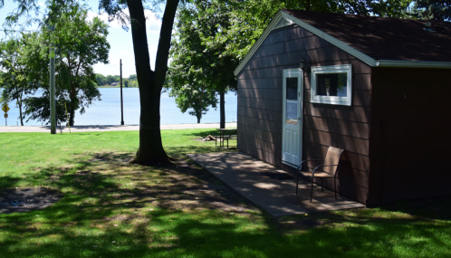 A small brown cabin by a lake, surrounded by green grass and trees, with a chair on the patio.