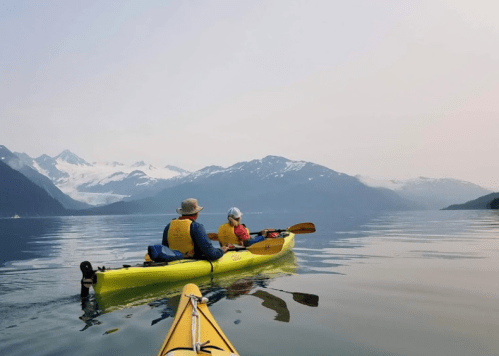 Two people in a yellow kayak on calm water, surrounded by mountains and a hazy sky.
