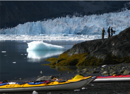 A serene landscape featuring kayaks on a rocky shore, with people observing a glacier in the background.