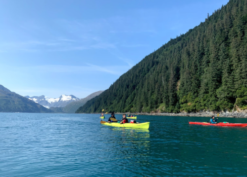 Two kayakers paddle in a serene blue lake surrounded by lush green mountains and distant snow-capped peaks.