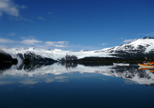 A serene landscape featuring a glacier, mountains, and calm waters reflecting the blue sky and clouds.