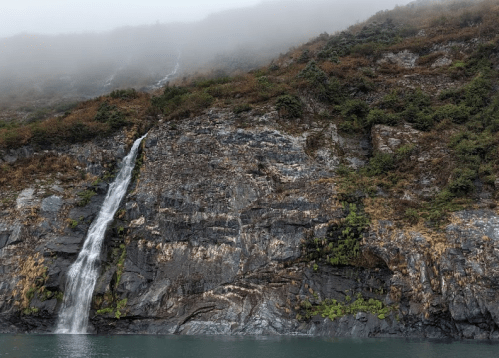 A waterfall cascades down rocky cliffs, surrounded by mist and greenery, reflecting in the calm water below.