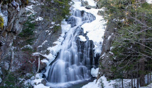 A cascading waterfall surrounded by snow-covered rocks and evergreen trees in a serene winter landscape.