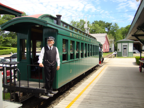 A vintage green train with a conductor standing on the platform at a quaint station surrounded by trees.