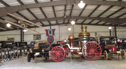 A vintage vehicle exhibit featuring classic cars, a steam engine, and an early airplane in a spacious indoor setting.