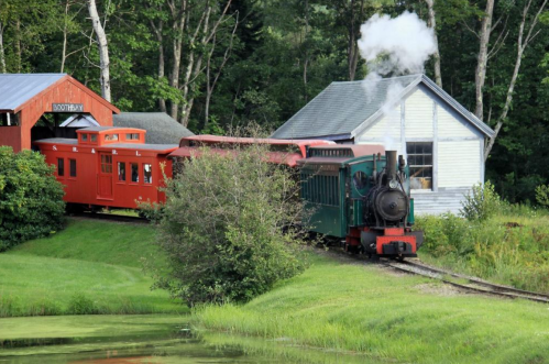A vintage steam train with red and green cars travels past a white building and lush greenery, emitting steam.