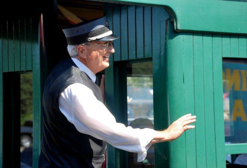 A smiling conductor in a vintage uniform stands by a green train car door, ready to assist passengers.