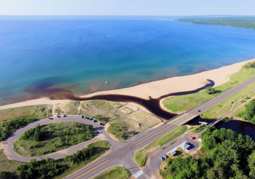 Aerial view of a sandy beach meeting a blue lake, with a winding stream and a road alongside lush greenery.