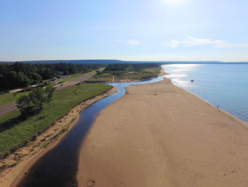 Aerial view of a sandy beach with a winding river, surrounded by greenery and a clear blue sky.
