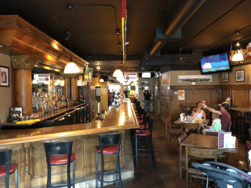 A spacious bar area with wooden accents, tables, and a TV, featuring patrons enjoying drinks and food.