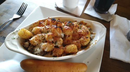A dish of cheesy tater tots topped with meat, served with a soft breadstick on the side.