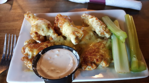 A plate of cheesy chicken wings with celery sticks and a side of dipping sauce.
