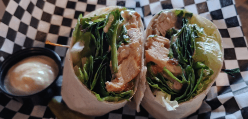 A close-up of a halved wrap filled with greens and chicken, served with a side of dipping sauce on a checkered plate.