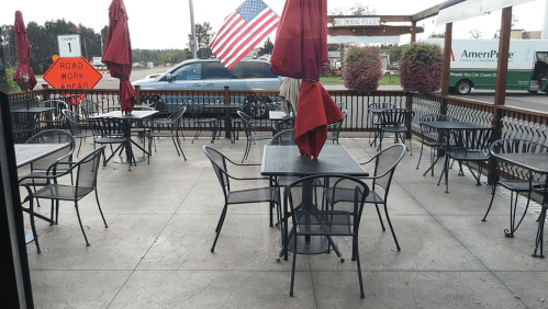 Empty outdoor dining area with black tables and chairs, red umbrellas, and an American flag in the background.