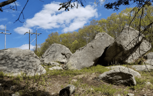 A rocky landscape with large boulders, surrounded by trees and power lines under a blue sky with fluffy clouds.
