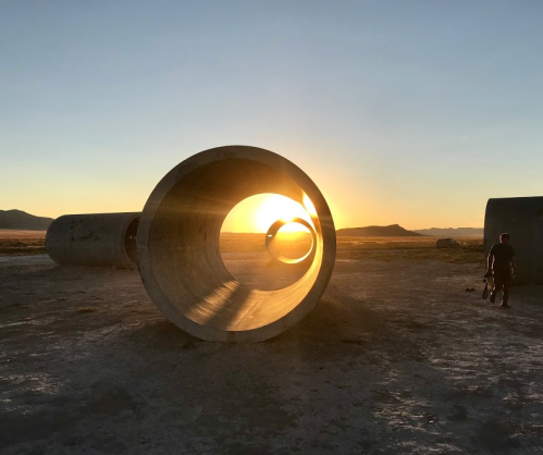A person stands near large concrete pipes at sunset, with the sun shining through one of the pipes in the foreground.